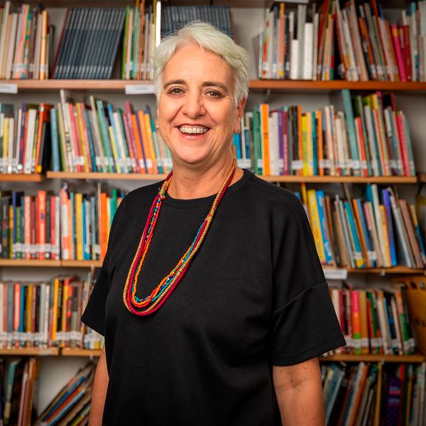 Foto de Roberta Panico, mulher de pele clara, cabelos brancos lisos e curtinhos. Ela posa sorridente na frente de uma estante repleta de livros.