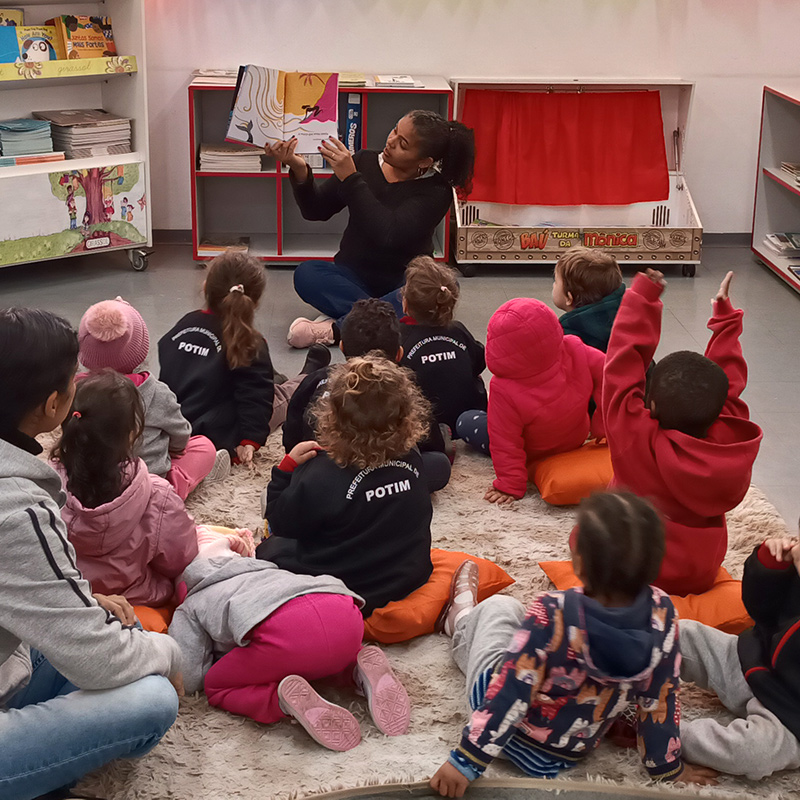 Foto de crianças diversas de costas, sentadas no chão e olhando para uma mulher que está na frente da sala. Ela mostra um livro para eles.