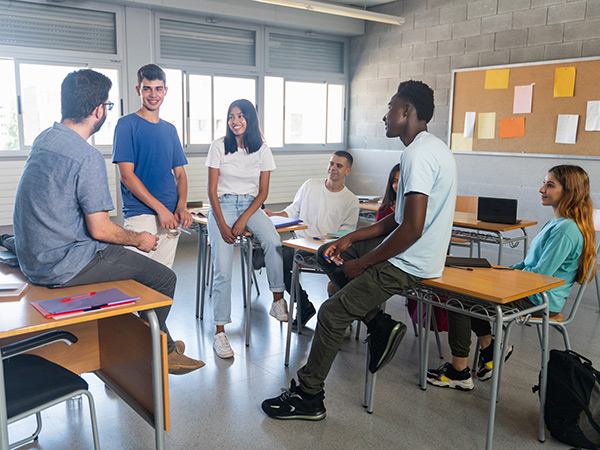 Foto de uma sala de aula com um professor e um grupo diverso de estudantes interagindo. Alguns estão sentados em carteiras escolares, outros sobre a mesa e um deles está em pé