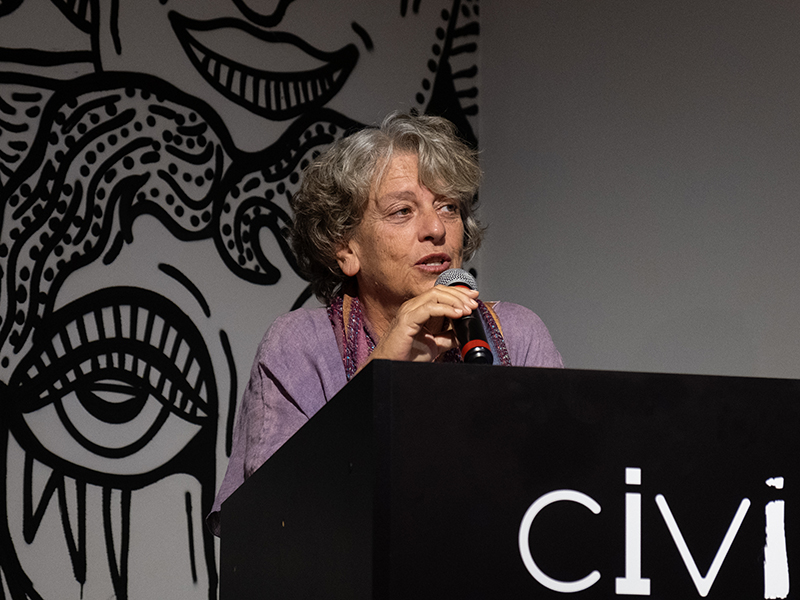 Foto de Ana Lima, uma mulher de pele branca e cabelos grisalhos na altura do queixo, com um microfone na mão na frente de um púlpito