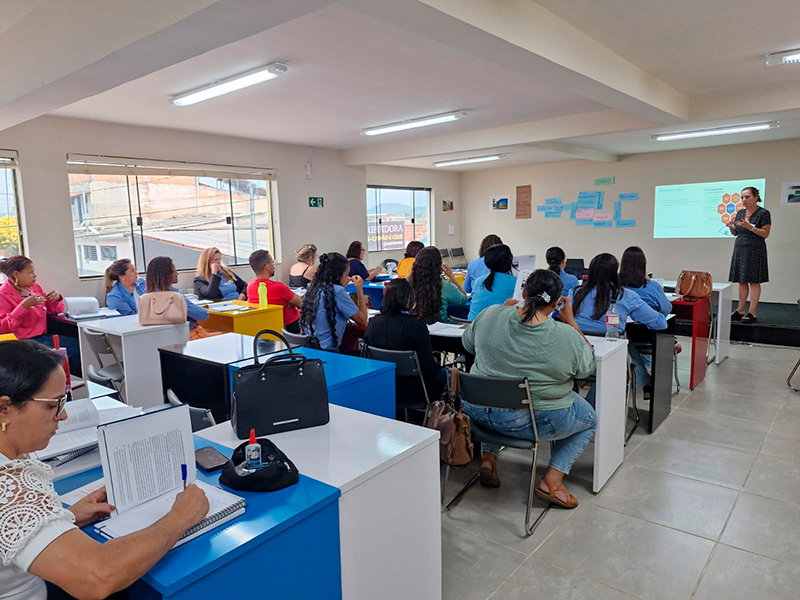 Foto tirada do fundo de uma sala de aula com um grupo diverso de pessoas acompanhando umaapresentação. Há uma pessoa em pé com uma projeção sendo feita na parede