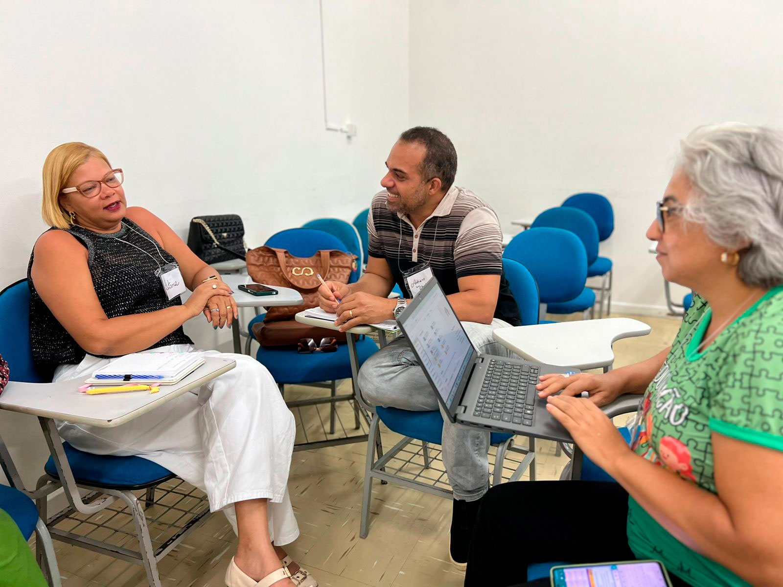 Foto com duas mulheres e um homem sentados em carteiras escolares em uma sala de aula. Eles se oham e conversam