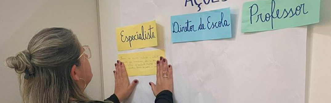Foto de uma mulher loira colando uma anotação em papel em um cartaz. Nele, há os textos: "Especialista, Diretor de escola e Professores".