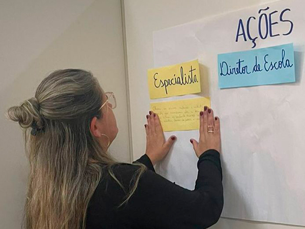 Foto de uma mulher loira colando uma anotação em papel em um cartaz. Nele, há os textos: 