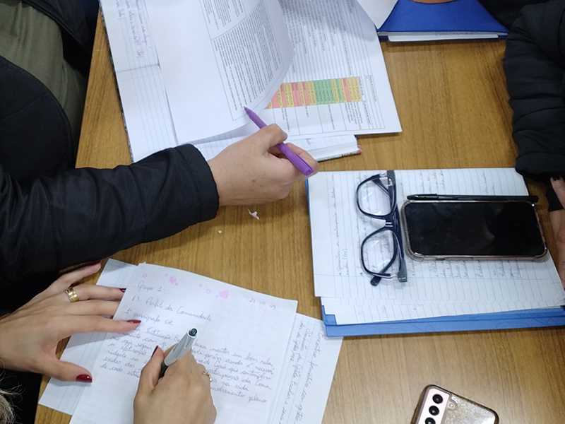 Foto em detalhe de uma mesa com papéis e cadernos sobre ela. Há algumas mãos escrevendo