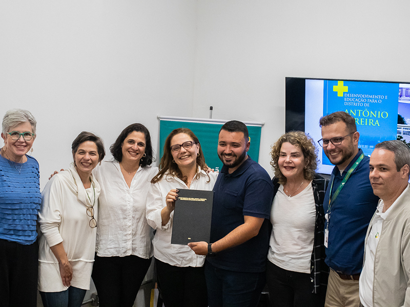 Foto de um grupo diverso de pessoas posando lado a lado. No centro da imagem, está um documento encadernado segurado por duas delas.