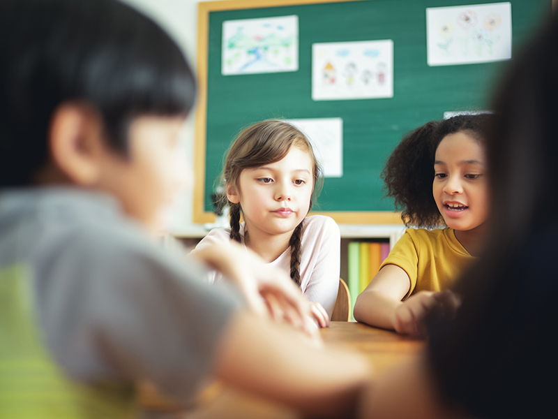 Foto de crianças diversas em uma sala de aula. A menina no centro faz uma careta como se estivesse descontente