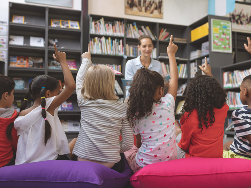 Foto de crianças diversas de costas com a mão levantada, Na frente delas, desfocada, há uma mulher segurando um livro na mão.