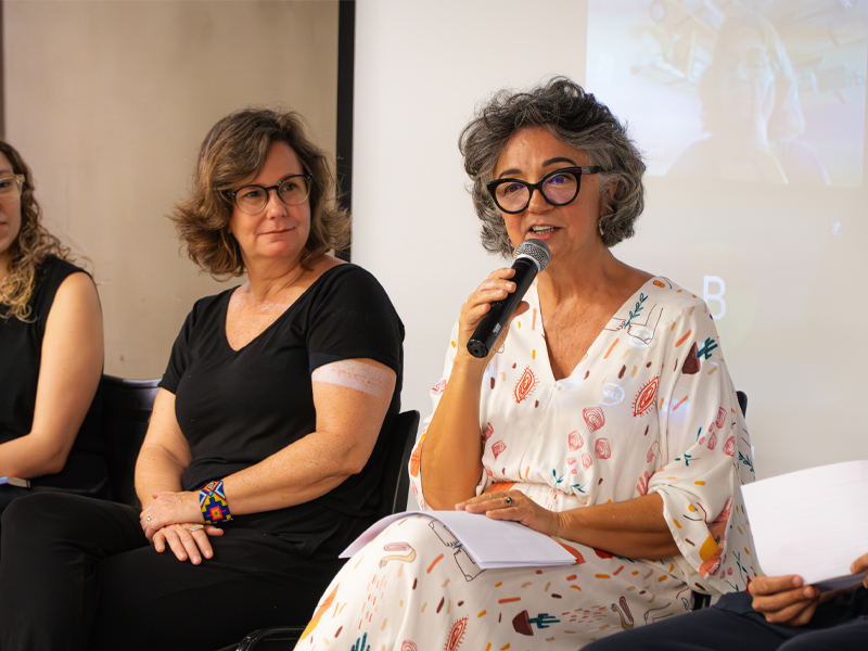 Foto de Tereza Perez sentada, ao lado de Juliana Ruschel, com um microfone na mão