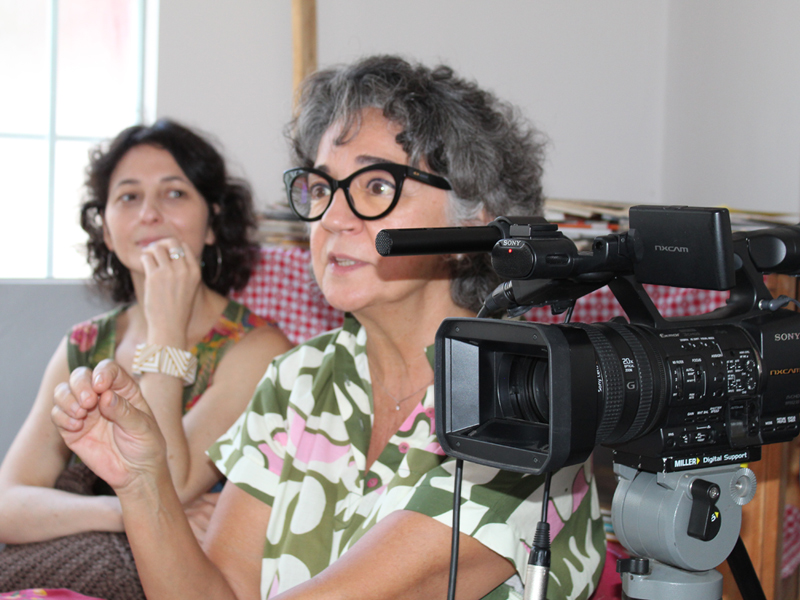 Foto de Tereza Perez gesticulando com uma filmadora ao seu lado. No fundo desfocado, está Carolina Glycerio