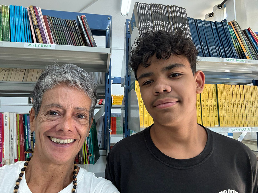 Foto de Vera, uma senhora de pele branca e cabelos grisalhos bem curtos, ao lado de Gustavo, um garoto de pele negra e cabelos curtos cacheados. Atrás deles há duas estantes repletas de livros.