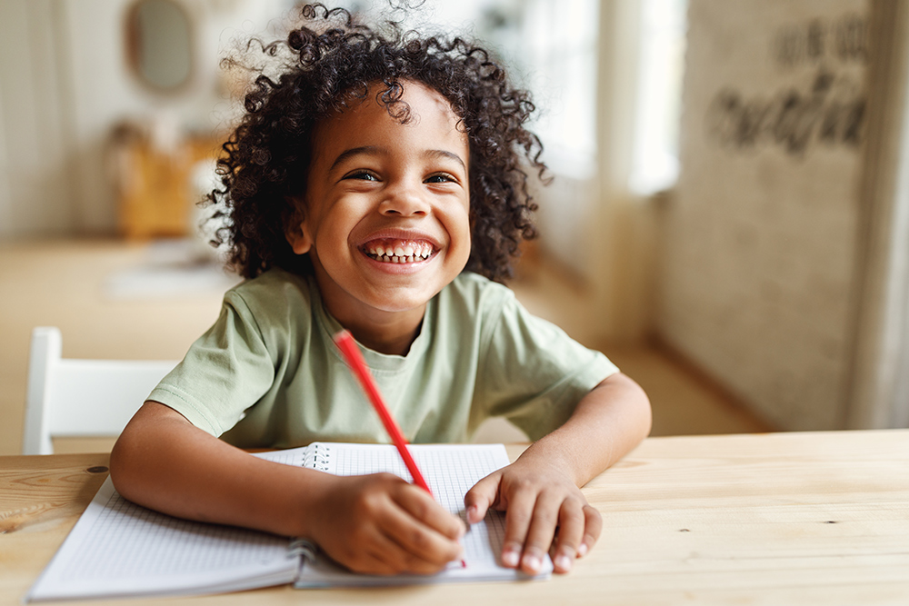 Foto de um menino de pele negra e cabelos cacheados sorrindo e com um lápis na mão, que está apoiado sobre um caderno em cima de uma mesa