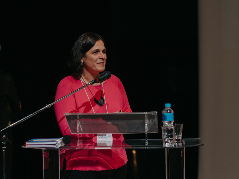 Foto de Patrícia Diaz, uma mulher de pele branca e cabelos lisos escuros, atrás de um púlpito com um microfone.