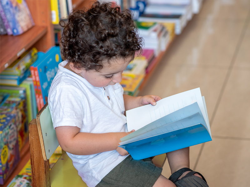 Foto de menino pequeno de lado sentado em uma cadeira olhando para um livro que está em suas mãos.