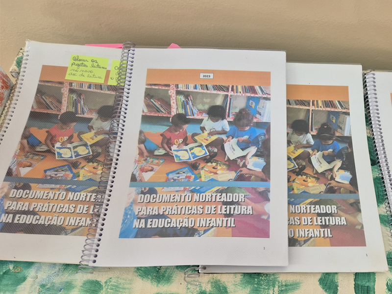 Foto de documentos encadernados sobre uma mesa. Na capa, há a foto de crianças e o texto "Documento norteador para práticas de leituras na Educação Infantil".