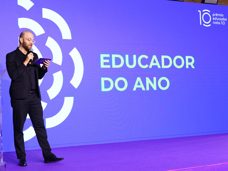 Foto de Helder Guastti, um homem de pele clara usando óculos de grau em pé e com um microfone na mão. Ao lado dele, há os dizeres "Educador do ano" em um telão.