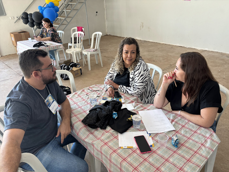Foto de um homem e duas mulheres ao redor de uma mesa. Sobre ela, há celulares e diversos papéis