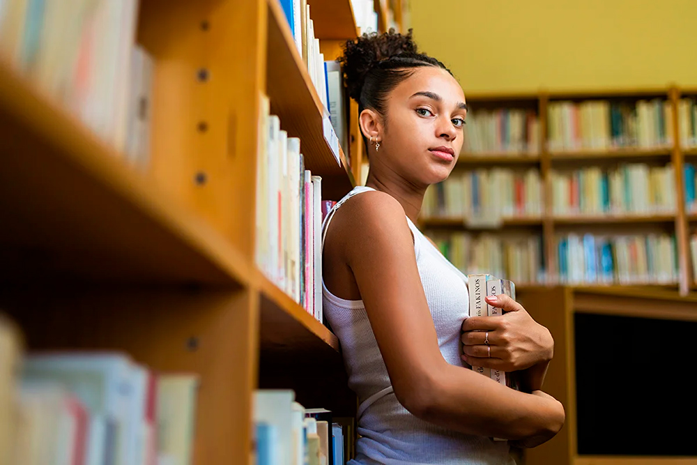 Foto de garota jovem negra de perfil encostada em uma estante de madeira cheia de livros. Ela segura dois livros nas mãos