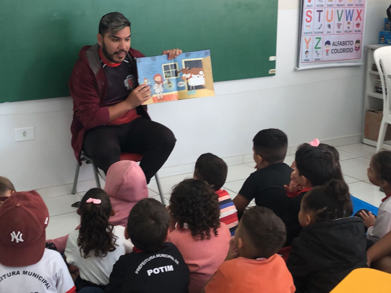 Foto de um homem sentado em uma cadeira na frente de uma sala de aula segurando um livro. No chão, há um grupo diverso de crianças olhando pra ele.