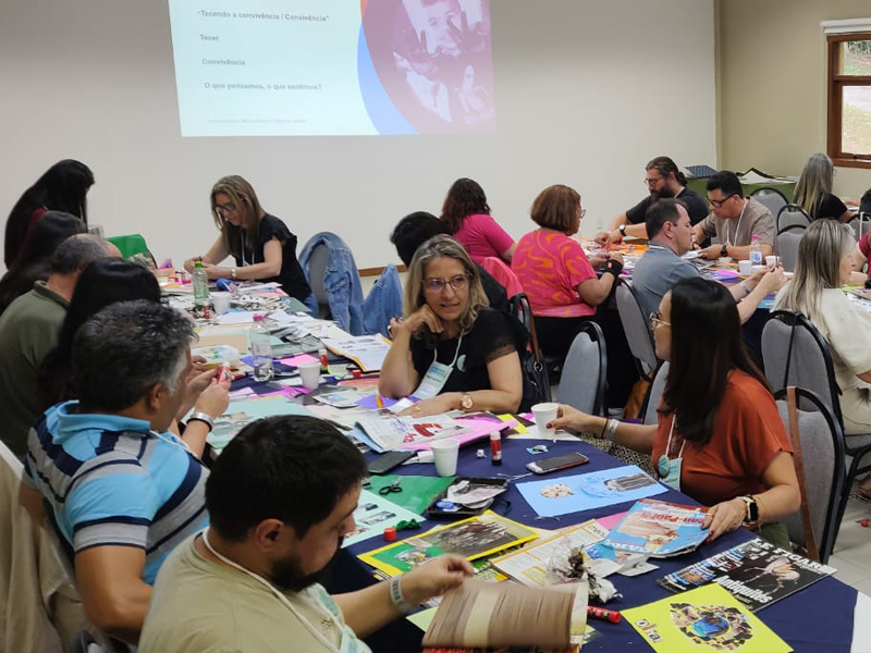 Foto de pessoas diversas em uma sala de aula. Há uma projeção em um telão e diversos objetos sobre as mesas, como revistas, colas e tesouras.