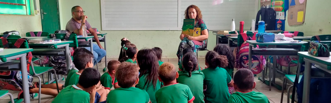 Foto de um grupo diverso de crianças, de costas pra imagem, sentadas no chão. Elas olham para uma mulher na frente da sala que segura um livro em suas mãos.