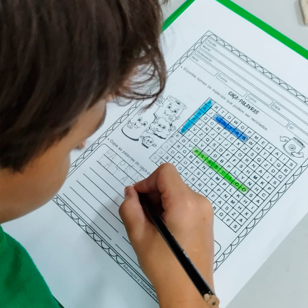 Foto em close e tirada de costas de um garoto sentado em uma carteira escolar escrevendo palavras em uma folha de atividade com um caça-palavras.