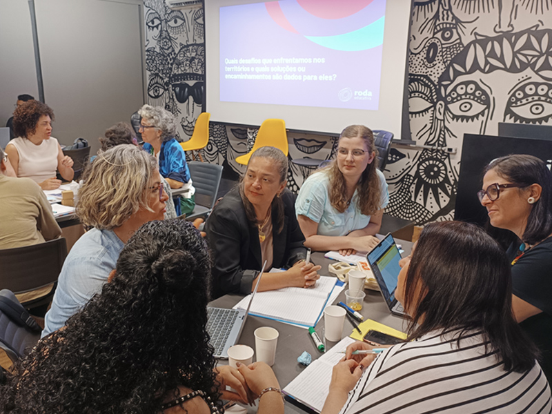 Foto de um grupo diverso de mulheres sentadas ao redor de uma mesa, olhando para uma delas, que está falando. Ao fundo, há um telão.