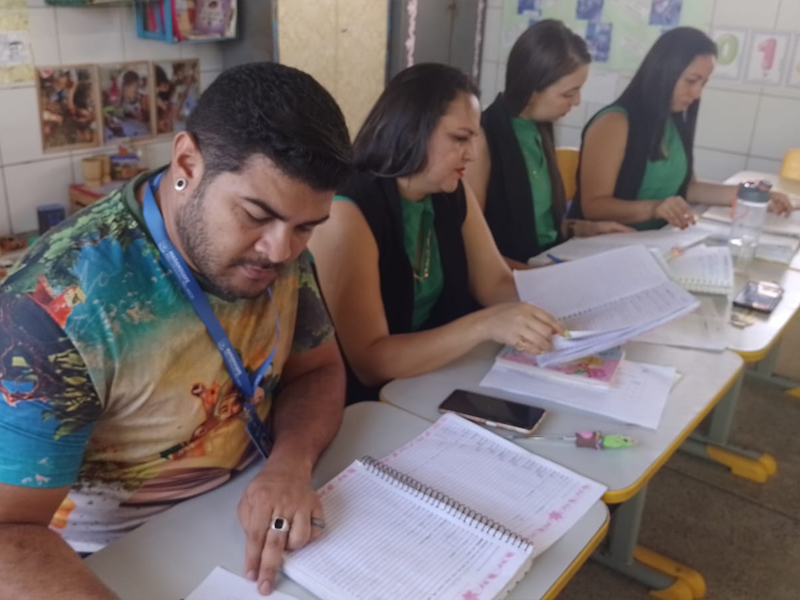 Foto de 4 pessoas, 1 homem e 3 mulheres, lado a lado, olhando para cadernos que estão na mesa à sua frente.