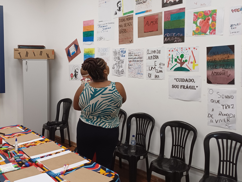 Foto de diversos cartazes estilo lambe-lambe colados em uma parede. Há uma mulher com um bebê no colo olhando para eles.