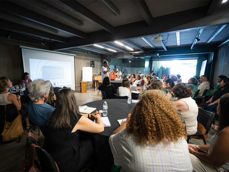 Foto de um homem em pé, na frente de um telão, com um microfone na mão. Há um grupo diverso de pessoas olhando para ele.