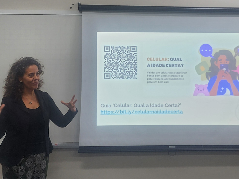 Foto de Helena Mendonça gesticulando ao lado de um telão. Na tela, há a projeção da divulgação do guia “Celular: qual a idade certa?”.