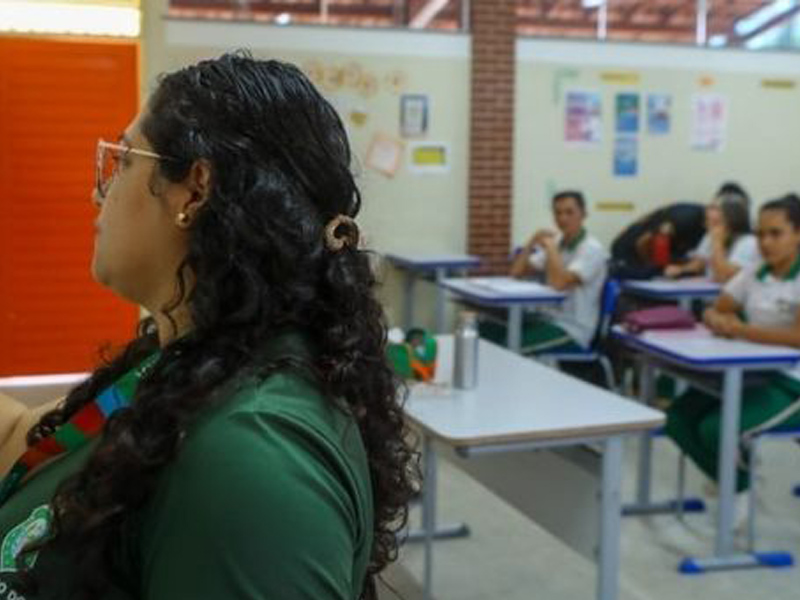 Foto de uma mulher em primeiro plano, no fundo desfocado, há adolescentes diversos em carteiras escolares.