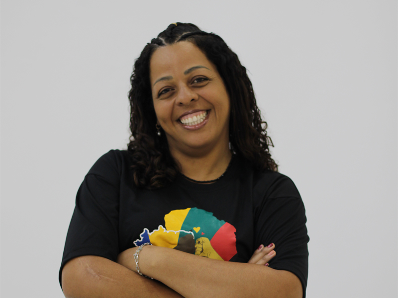 Foto de Juliana Ramos, uma mulher negra com tranças no cabelo que vai até a altura dos ombros. Ela está com os braços cruzados e sorrindo.
