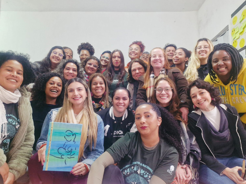 Foto de um grupo diverso de pessoas, em sua maioria mulheres, posando juntos e sorrindo, Umas delas segura um livro nas mãos com o desenho da silhueta de uma mulher amamentando um bebê.