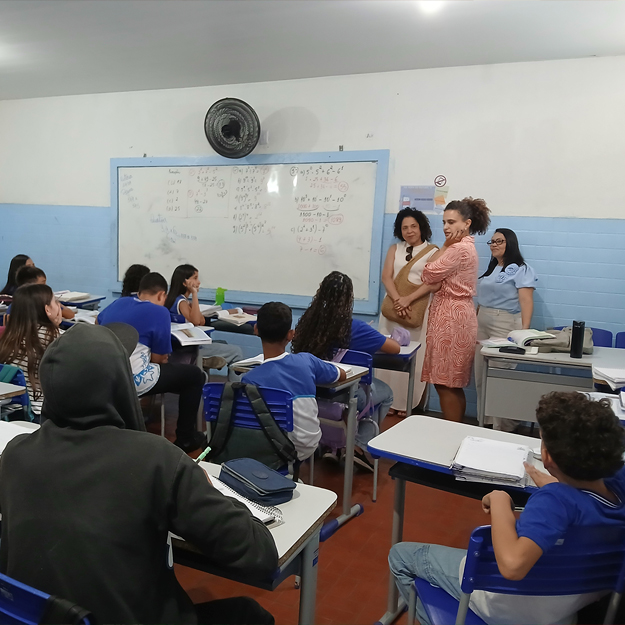 Foto de crianças diversas em uma sala de aula sentadas em carteiras escolares. Há três mulheres em pé na frente da sala.