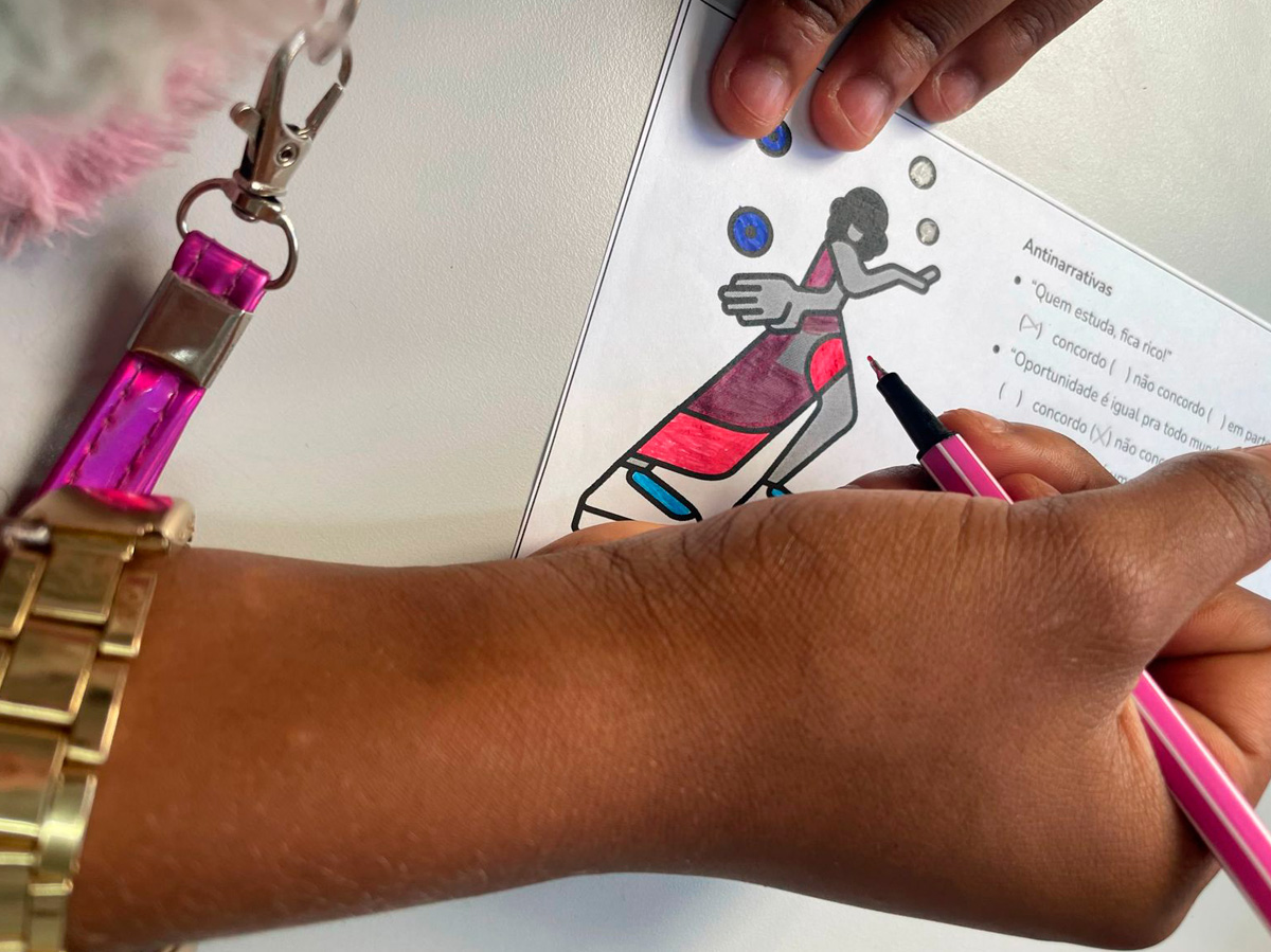 Foto em close de mão negra segurando uma caneta sobre um papel com o desenho de uma mulher e parte em texto para ser assinalada.