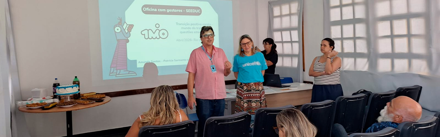 Foto de uma sala com 1 homem e 3 mulheres na frente fazendo uma apresentação ao lado de um telão. Nele á um apresentação com o logo da iniciativa 1MiO.