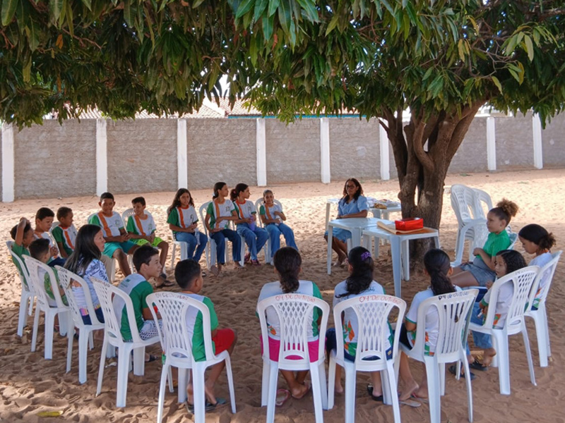 Foto de uma mulher adulta e um grupo diverso de crianças sentadas em cadeiras plásticas dispostas em formato de círculo ao redor de uma árvore em um ambiente externe de chão de areia.