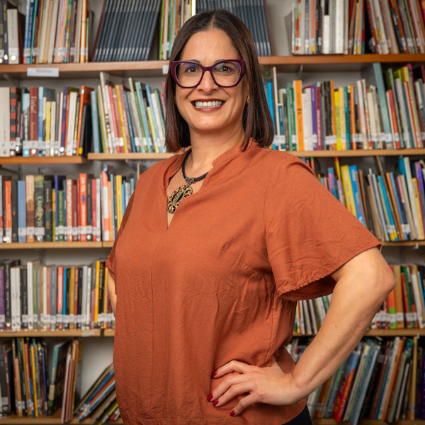 Foto de Patrícia Diaz, mulher de pele clara e cabelos castanhos escuros e lisos usando um colar com um grande pingente e óculos de grau com uma armação escura. Ela posa sorridente na frente de uma estante repleta de livros.