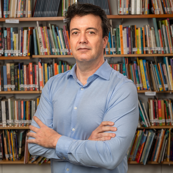 Foto de Ricardo Vilela, homem de pele branca e cabelos castanhos escuros. Ele veste camisa social e está de braços cruzados na frente de uma estante repleta de livros.