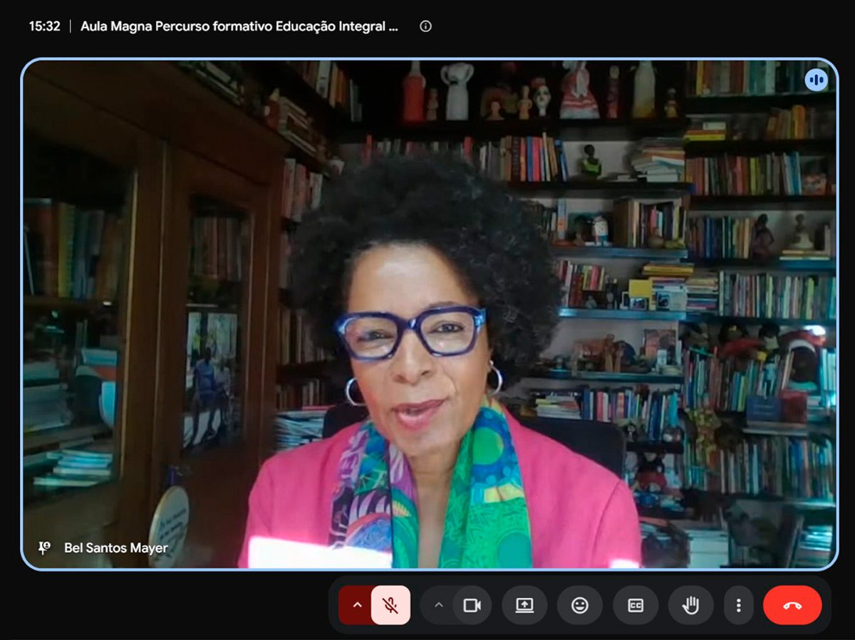 Print da gravação de uma aula magna feita pelo Google meet com Bel Santos Mayer na tela. Ela é uma mulher de pele negra, com cabelos curtos e volumosos, que usa óculos de grau com armação escura e lenço colorido ao redor do pescoço.