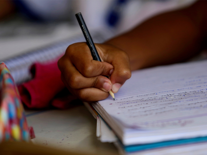 Foto da mão de uma criança escrevendo com um lápis em um caderno.