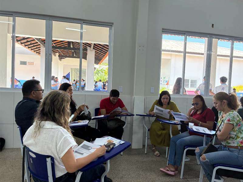 Foto de pessoas adultas diversas em uma sala olhando para materiais impressos diversos que estão em suas mãos ou sobre as mesas. Atrás delas, pela janela, há alguns jovens em um pátio.